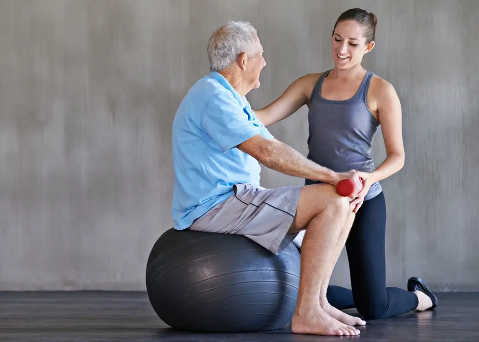Senior sitzt auf einem Balanceball, unterstützt von einer Therapeutin – gezielte Krankengymnastik zur Förderung von Gleichgewicht, Stabilität und Körperkontrolle.